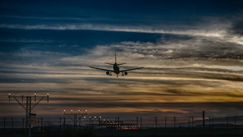 Avião visto de trás sobrevoando estruturas de energia, indo em direção do céu com nuvens e nas cores azul e alaranjado