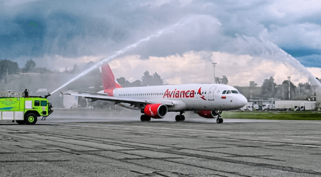 Avião branco com logomarca e nome da empresa Avianca em vermelho com jatos de água sendo jogados sobre avião parado na pista