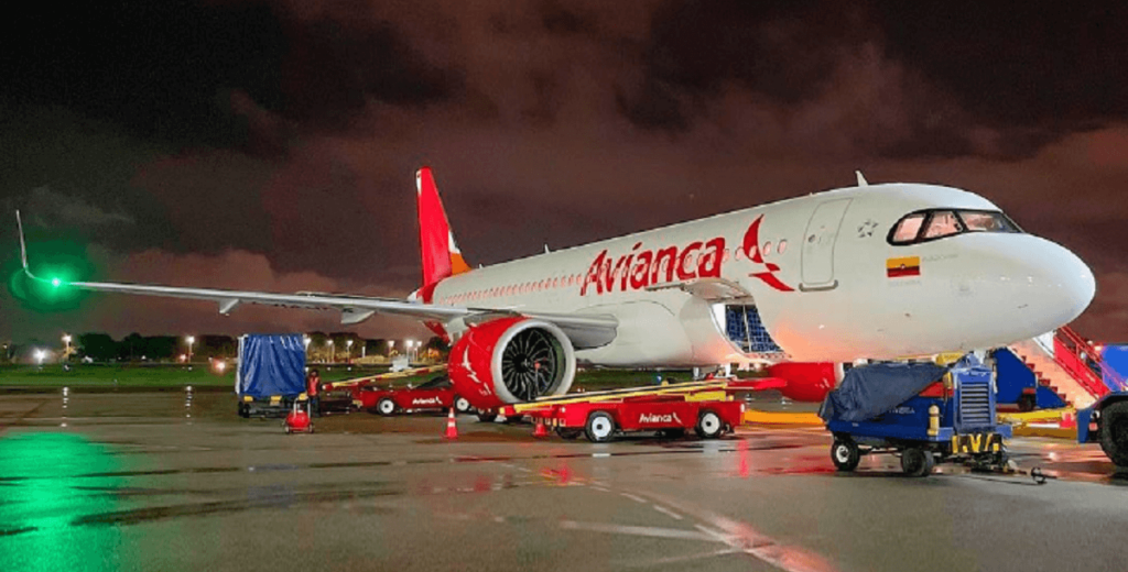 Avião branco da Avianca com logo e nome da empresa em vermelho em um aeroporto durante a noite