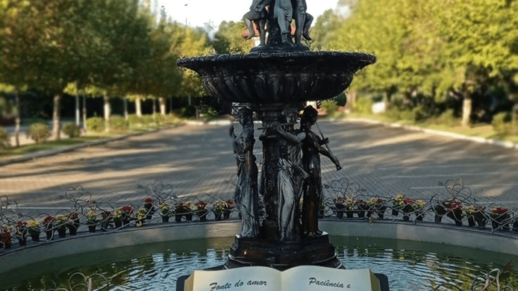 fonte em meio a praça, com flores na borda da fonte, um dia ensolarado