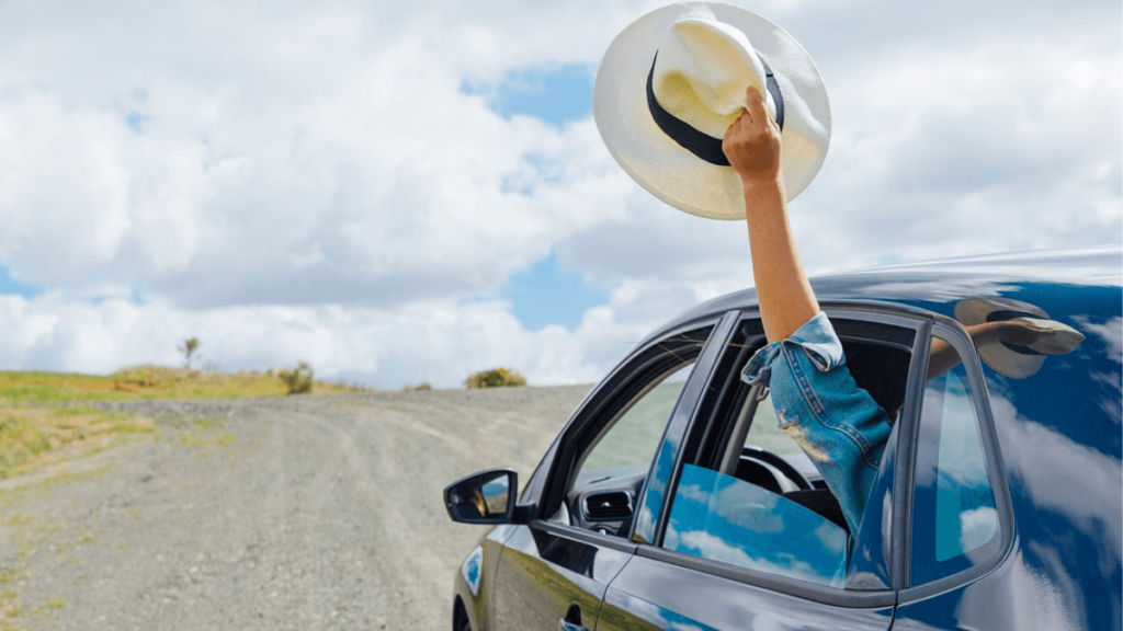 mulher viajando de carro preto com o braço estendido para fora segurando um chapéu branco