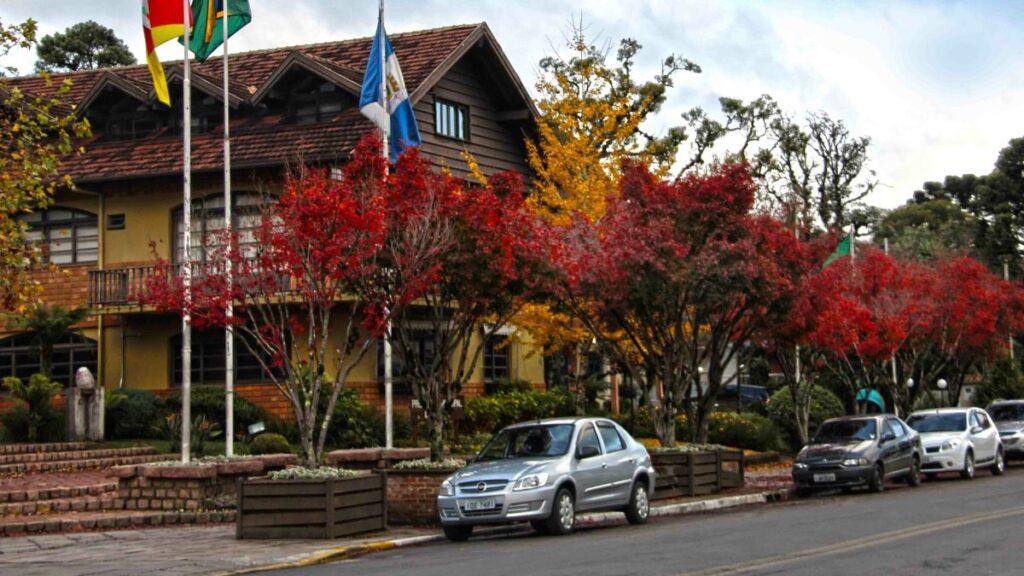Gramado Rio Grande do Sul com três bandeiras e alguns carros com uma casa de madeira ao fundo.