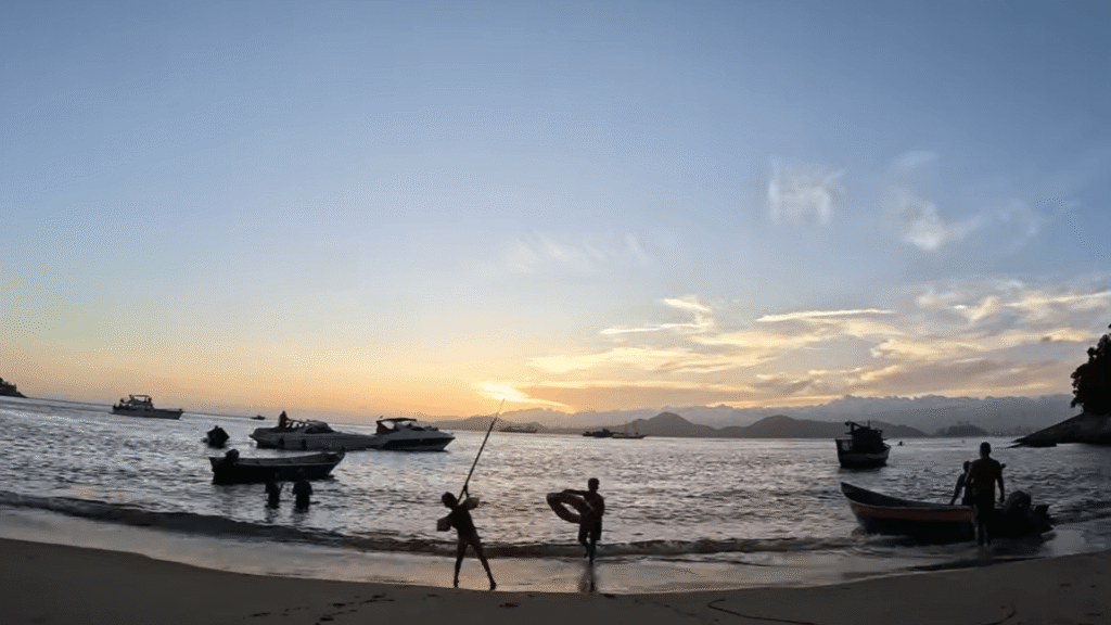 Praia do Sangava com pessoas brincando, barcos no mar e sol se pondo.
