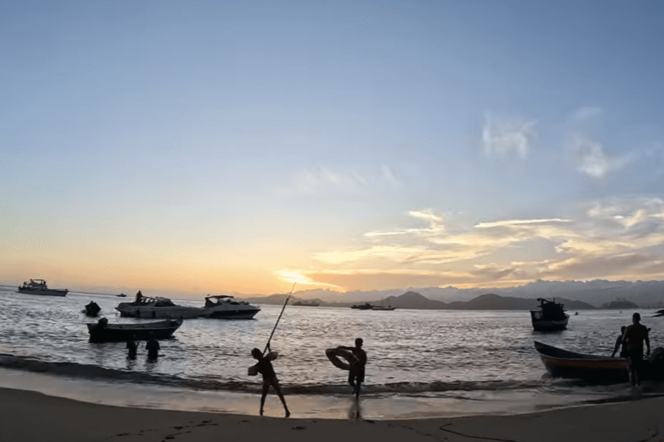 Praia do Sangava com pessoas brincando, barcos no mar e sol se pondo.