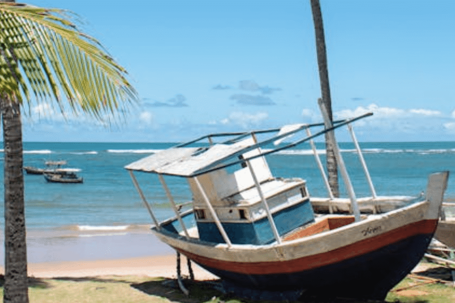 Praia de Camaçari de dia com um barco em cima da areia e ondas no mar com agua verde.